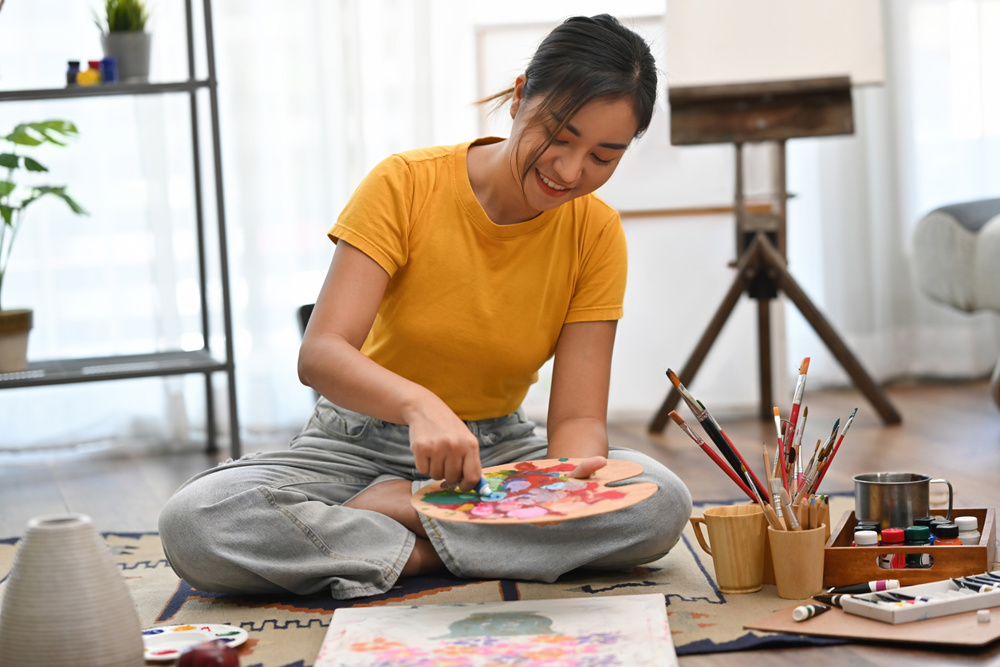 A young Asian woman in a yellow shirt sits on the floor, joyfully mixing paints on a wooden palette, surrounded by brushes, paint bottles, and an easel, ready to create her next masterpiece